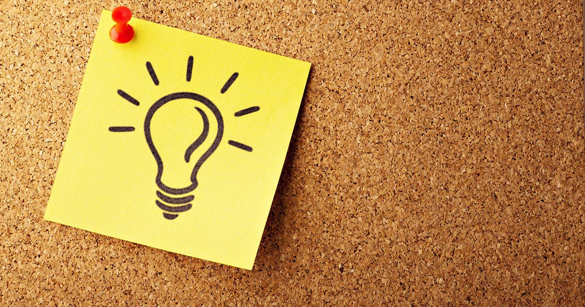 A yellow sticky note thumbtacked to a cork board with a drawing of a lightbulb on it.