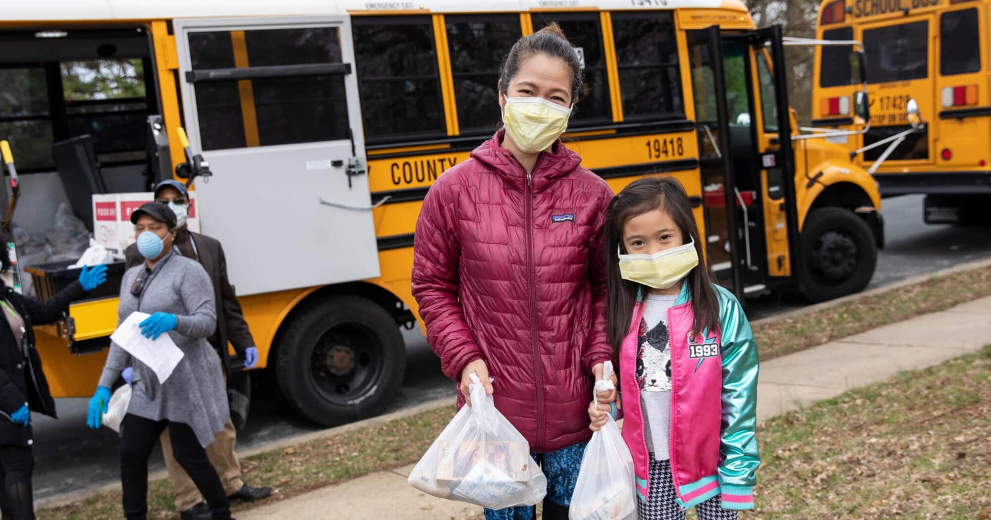 Image of a woman and a young girl with masks posing for a photo in front of a school bus holding plastic bags of food distributed to them by No Kid Hungry
