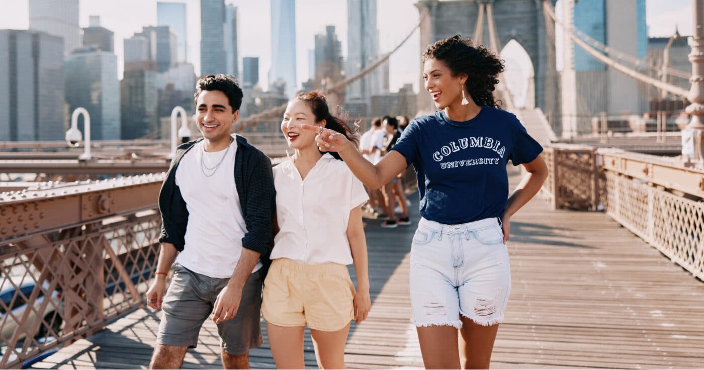 Three Columbia University students walk across a bridge with the New York City skyline pictured in the background.
