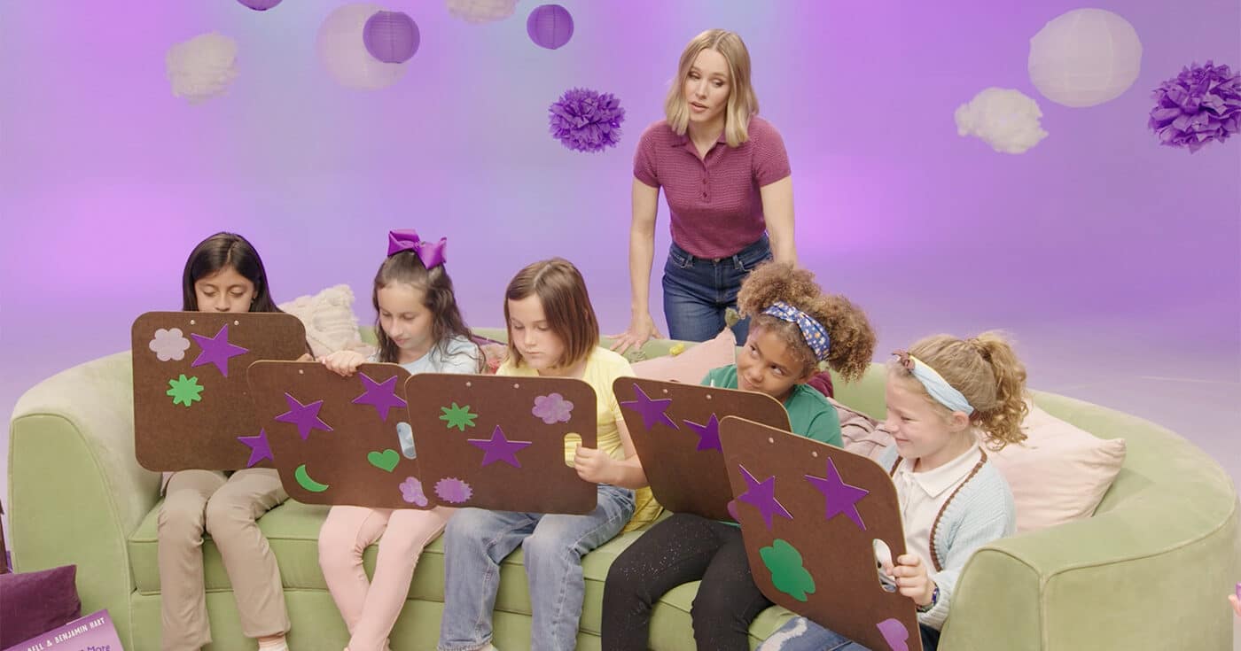 Kristen Bell instructs a couch full of girl scouts who are completing a worksheet on large clipboards.