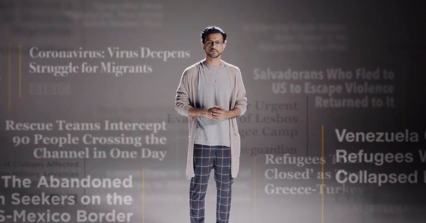 A man stands in front of a background of news headlines about the challenges refugees and migrants face.