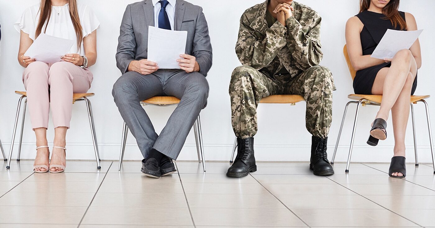 A series of people waiting in a waiting room. One of them stands out because they are wearing army fatigues.
