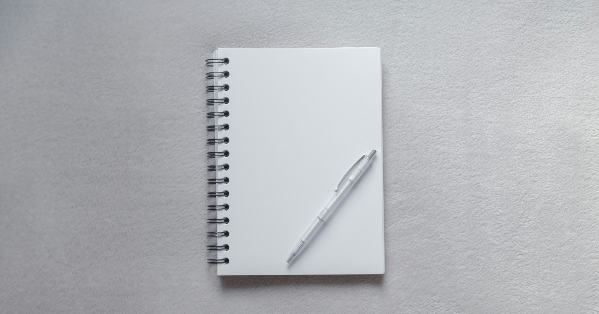 A spiral-bound blank notebook lies open on a light grey surface, with a clear pen resting diagonally on the empty page.