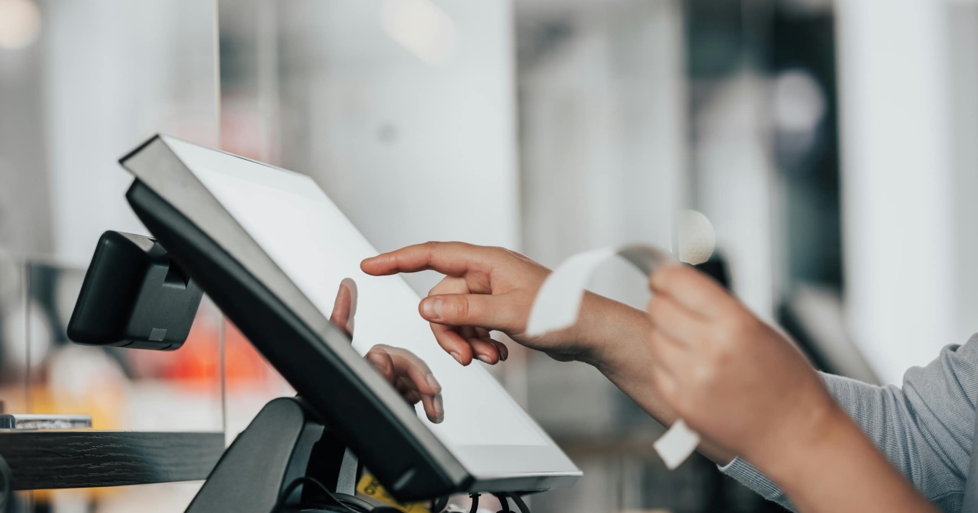 A person uses a touchscreen point-of-sale terminal while holding a paper receipt, possibly reviewing enrollment campaign questions, with a blurred background indicating an indoor setting.